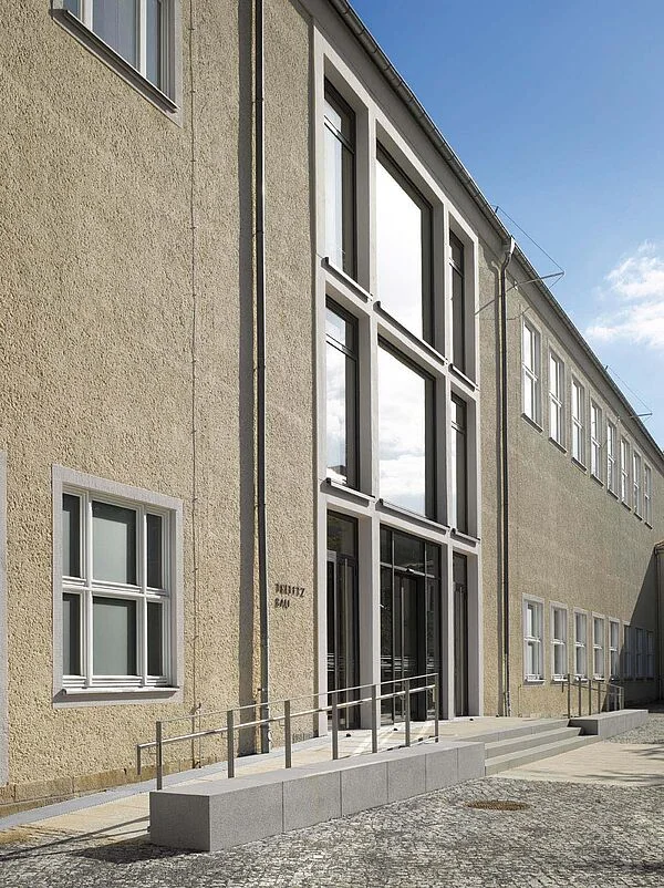 Doppelte Rampe mit jeweils einem Handlauf – TU-Dresden, Umbau und Modernisierung Hörsaalgebäude Trefftz-Bau. Die gegenüber dem Eingang angeordnete Treppe wird seitlich durch die Aufkantungen der Rampen begrenzt.